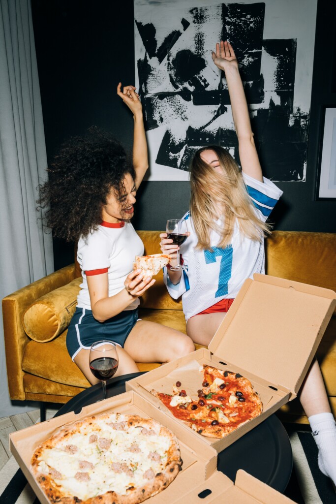 Zwei Freundinnen betreiben Selfcare indem sie Pizza essen und Wein trinken